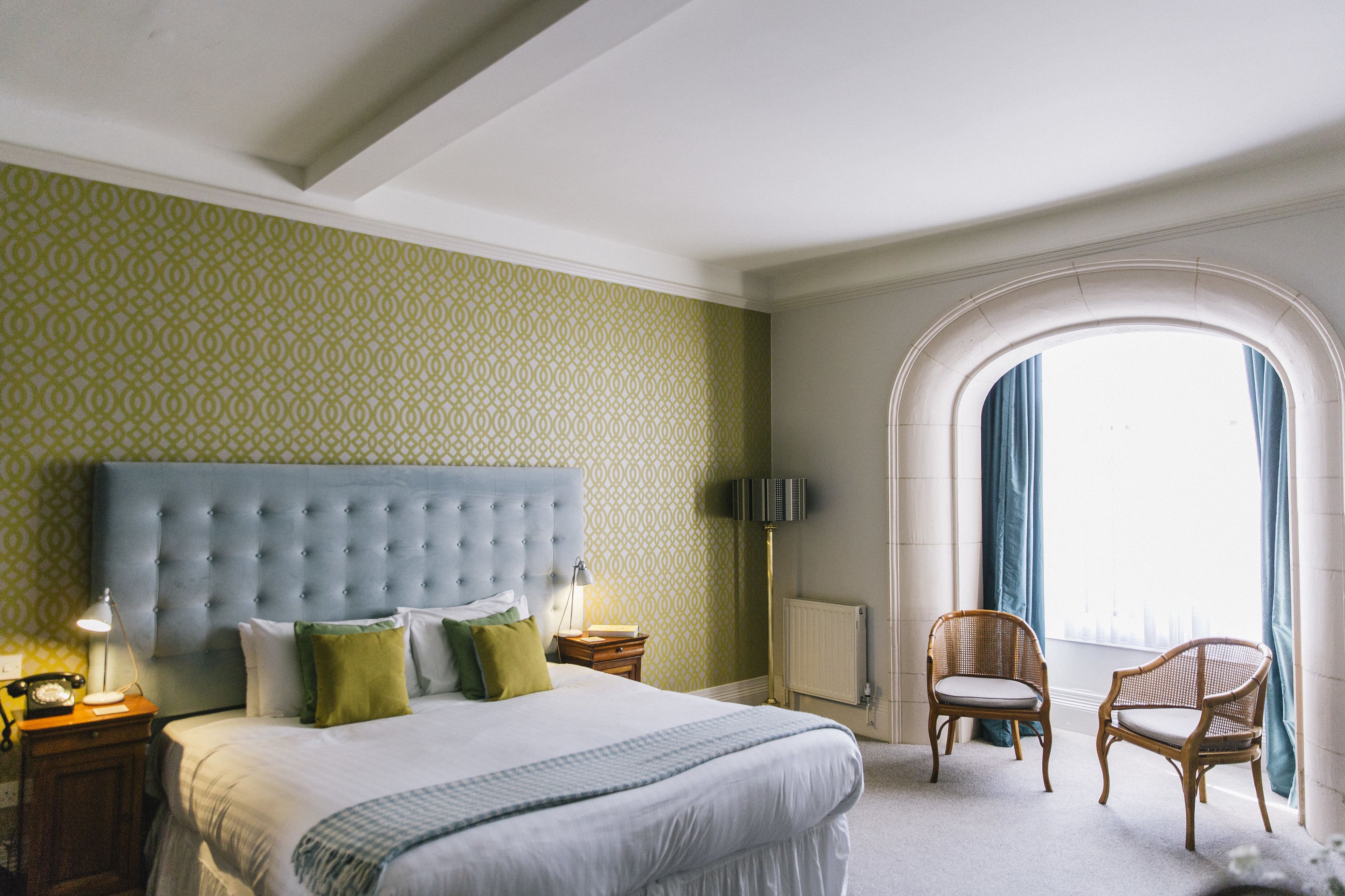 Habitación de hotel con una cama grande, cabecera capitonada, dos mesitas de noche, una lámpara, una silla y un ventanal de arco con cortinas.