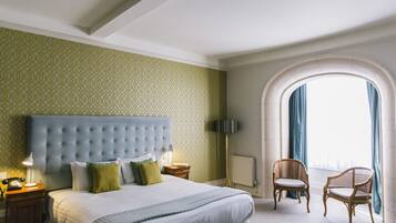 Habitación de hotel con una cama grande, cabecera capitonada, dos mesitas de noche, una lámpara, una silla y un ventanal de arco con cortinas.