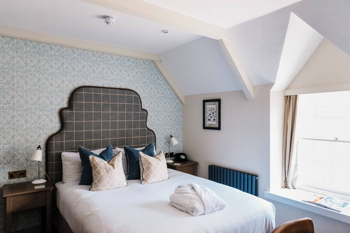 Une chambre à coucher avec un grand lit, un papier peint à motifs, un radiateur, une table de chevet avec une lampe et une fenêtre avec des rideaux.