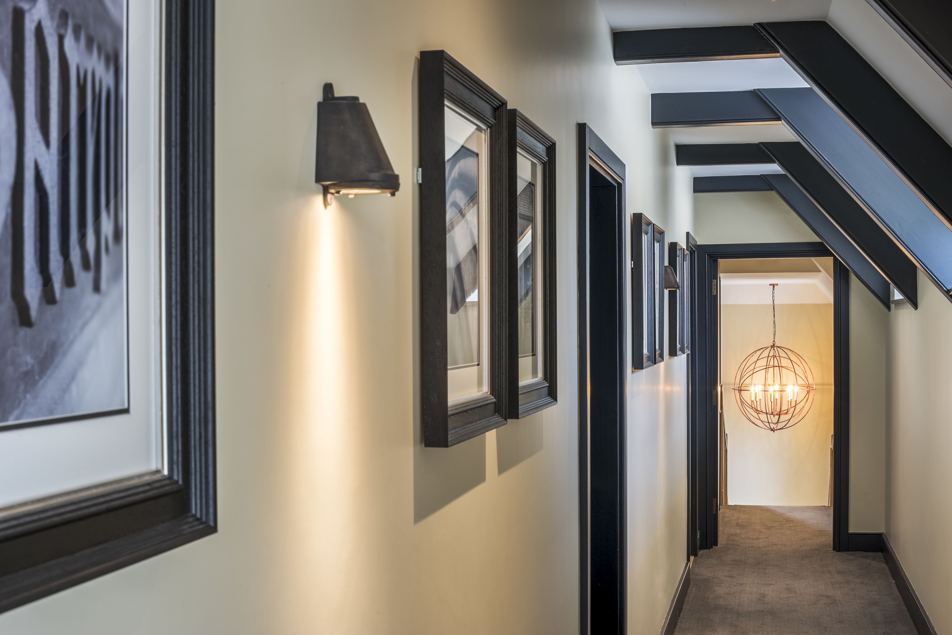 A hallway with framed artwork on the walls, a staircase with black railings, and a chandelier at the end.