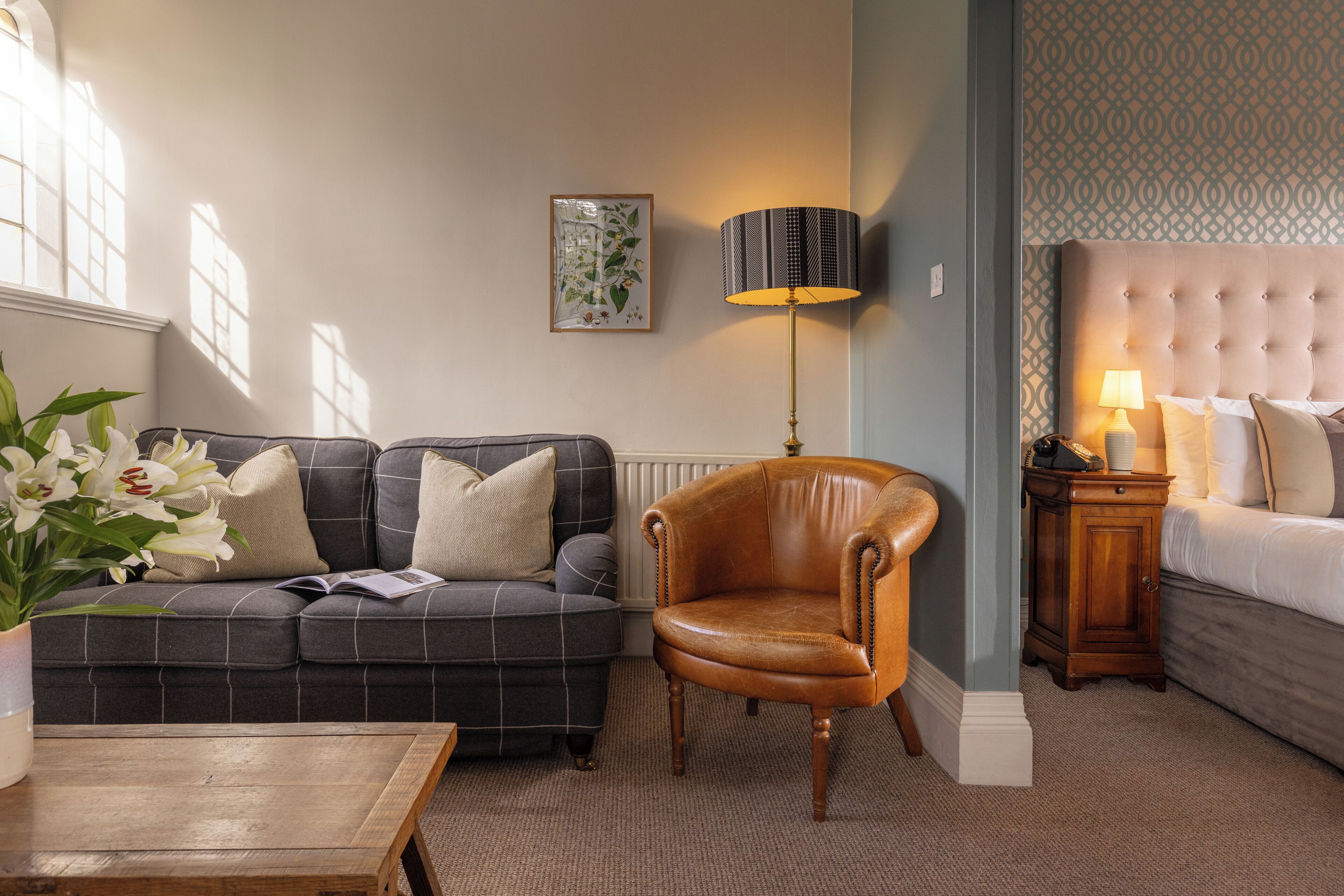 A cozy living room with a dark checkered sofa, a leather armchair, a wooden coffee table, and a bed with a tufted headboard.