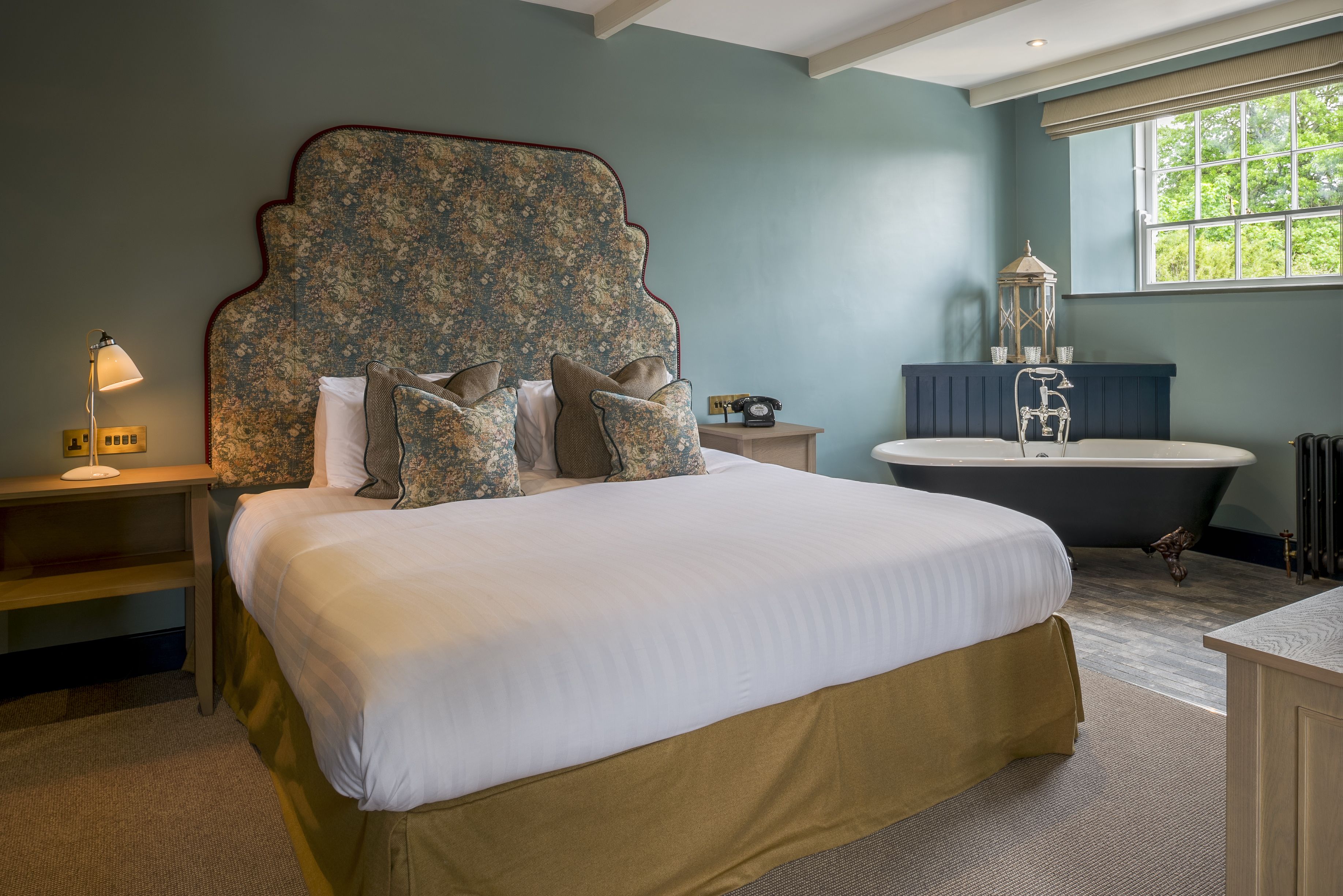 A bedroom with a large bed, a vintage telephone, a bathtub, and a window with a view of greenery.