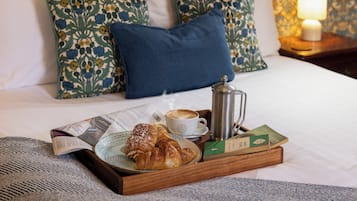 A breakfast tray with croissants, a cup of coffee, a newspaper, and a book on a bed with floral pillows and a nightstand with a lamp.