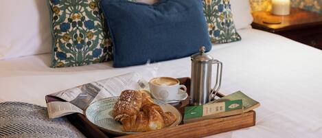 Bandeja de desayuno con croissants, una taza de café, un periódico y un libro sobre una cama con almohadas estampadas y una mesita de noche con una lámpara.