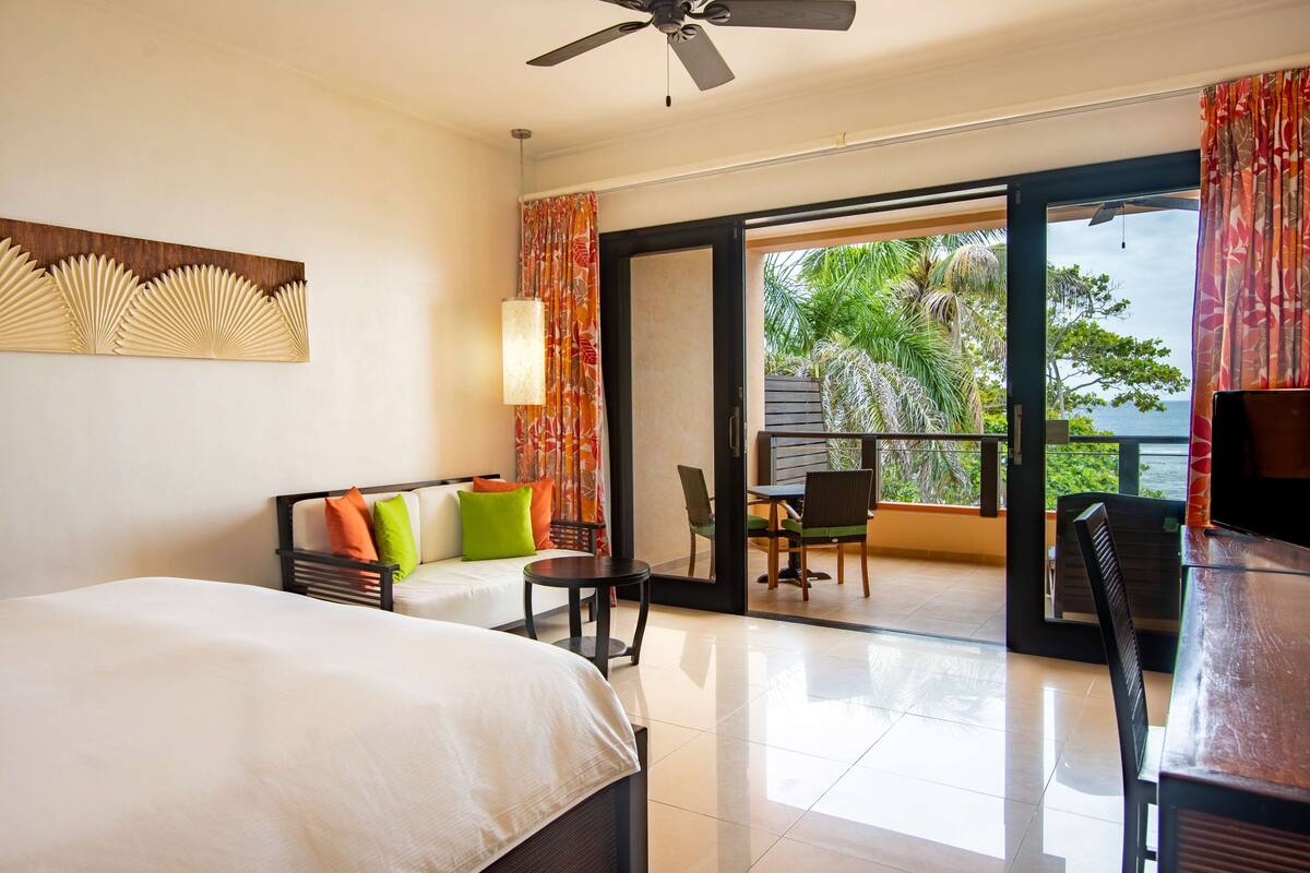 A hotel room with a bed, a sofa with colorful cushions, a small table, and a balcony with a view of the sea.