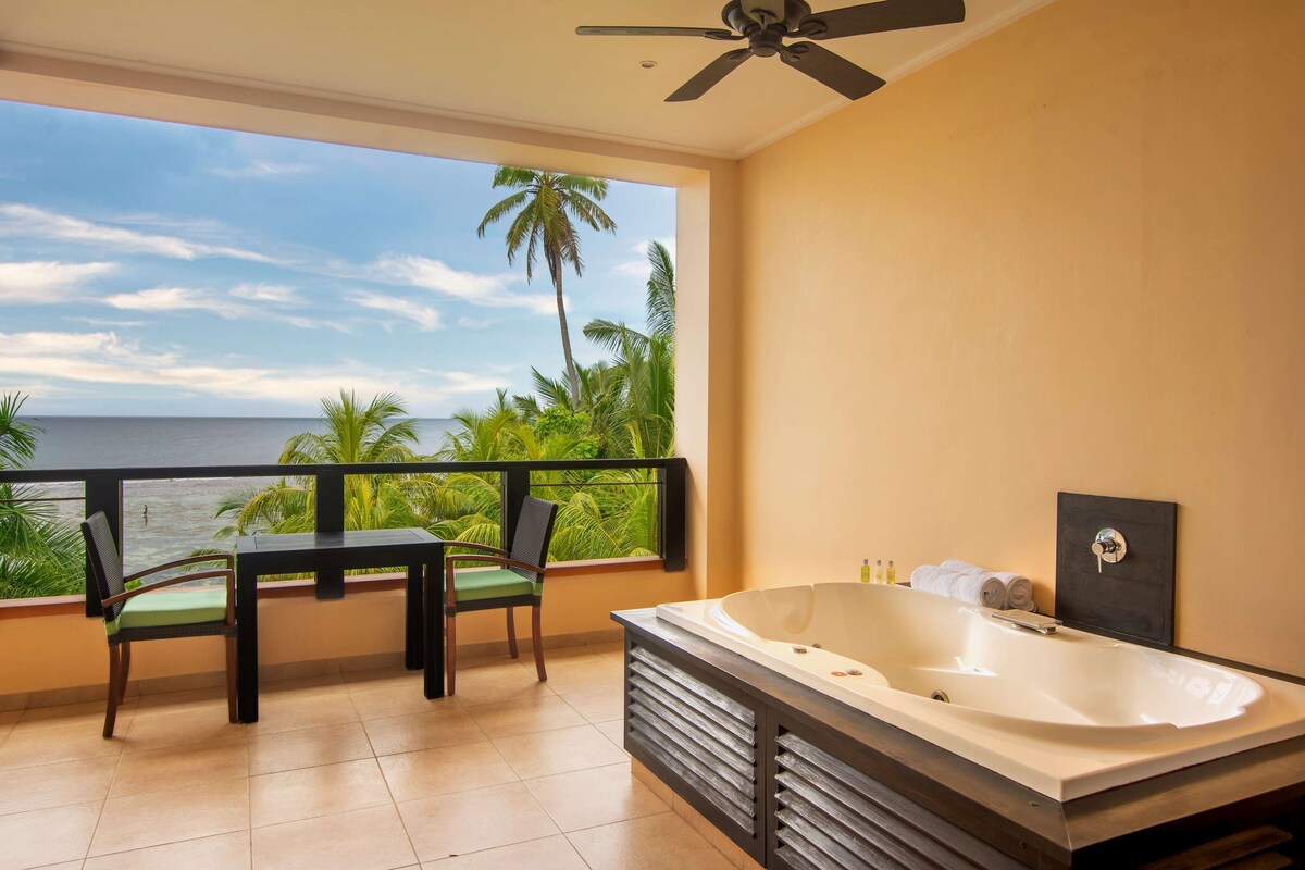 A balcony with a view of the ocean, a jacuzzi, and a table with chairs.
