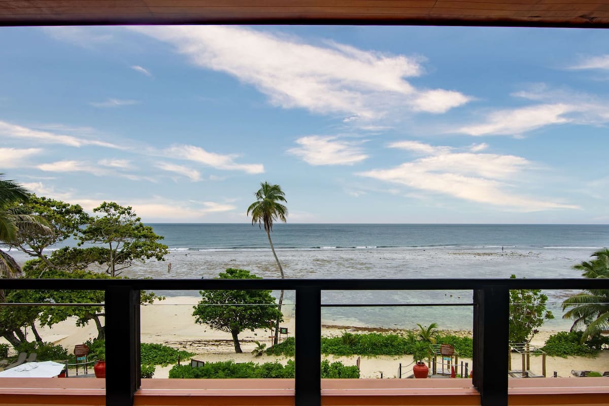 A beach view from a balcony with palm trees and a sandy beach.