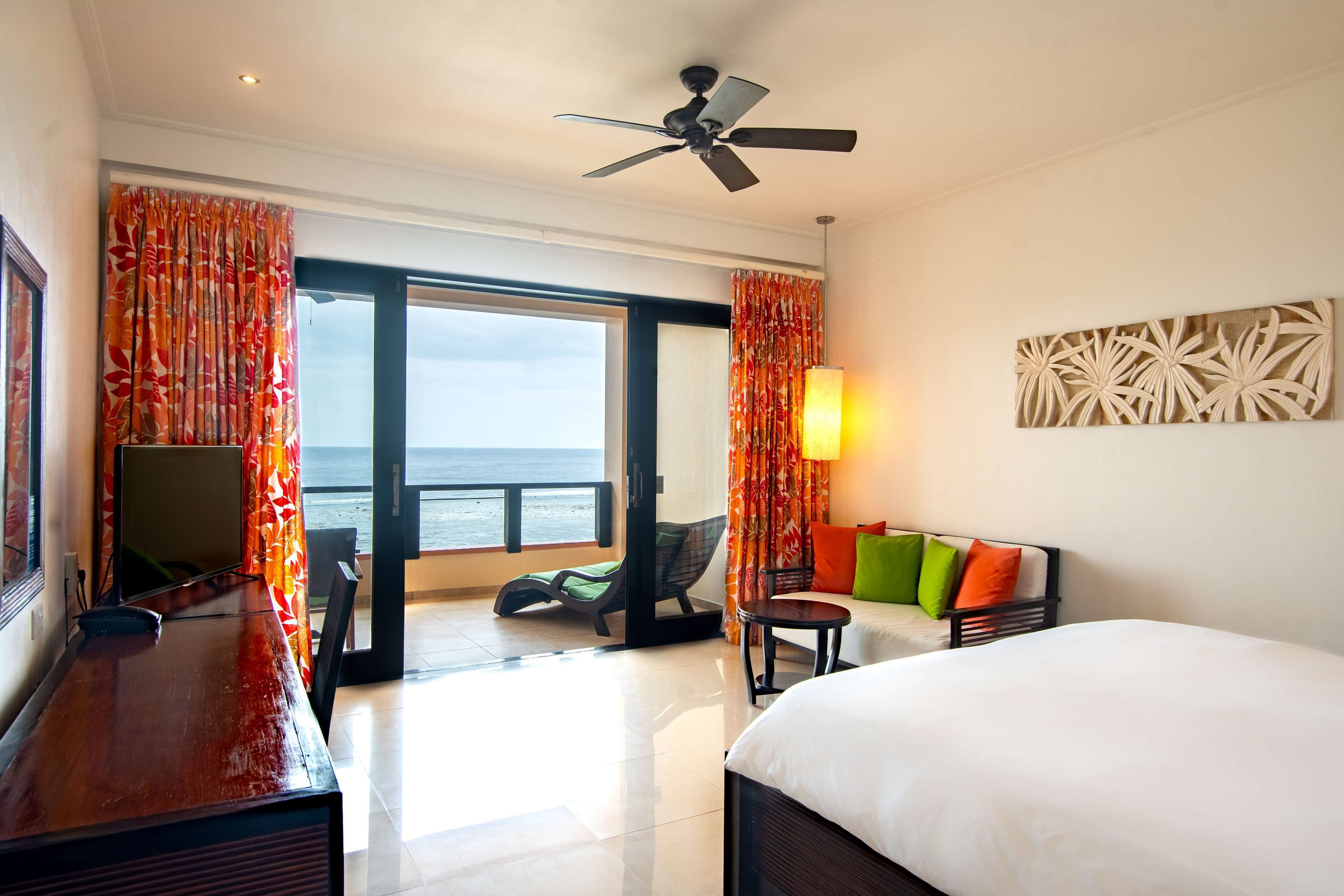 Quarto de hotel com cama, escrivaninha, poltrona, sofá com almofadas coloridas, ventilador de teto e vista para o oceano através da porta de vidro deslizante.