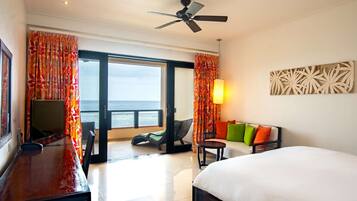 Quarto de hotel com cama, escrivaninha, poltrona, sofá com almofadas coloridas, ventilador de teto e vista para o oceano através da porta de vidro deslizante.