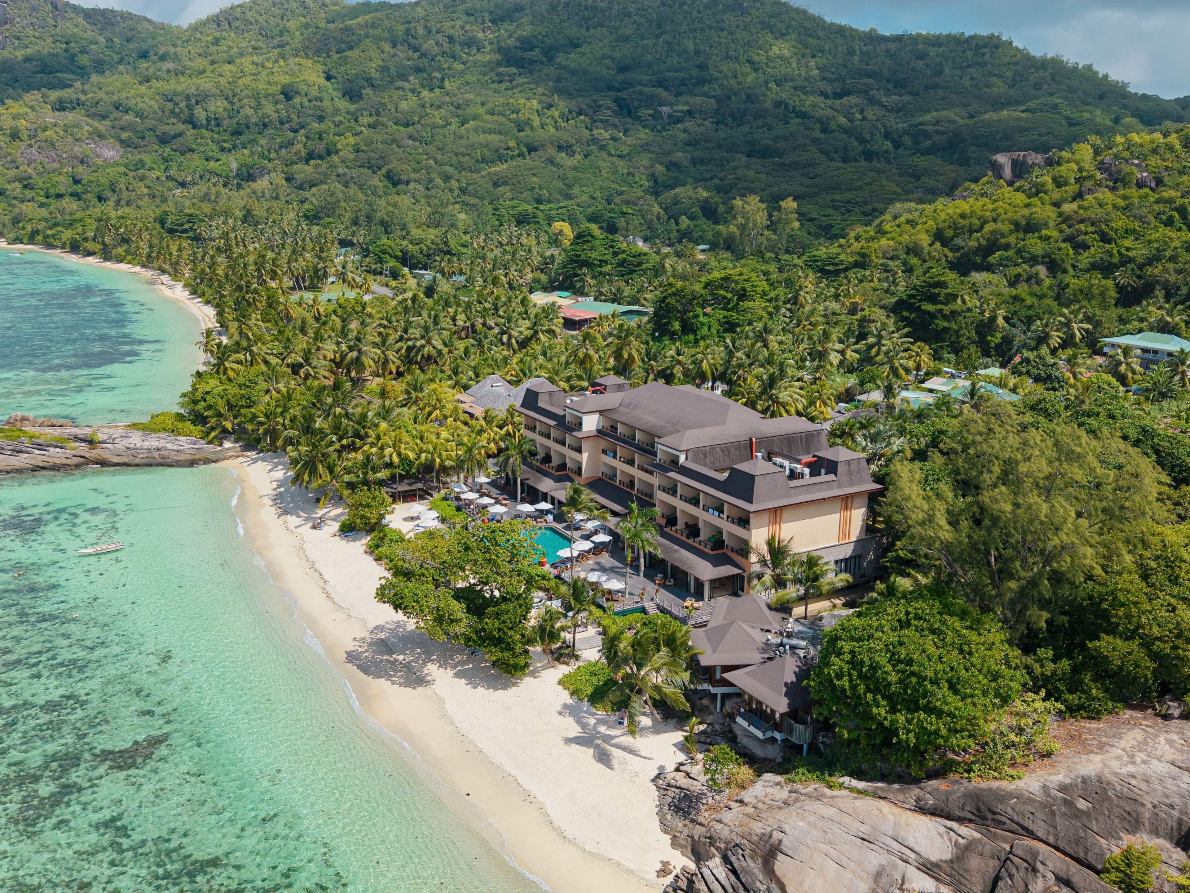 Un hotel frente a la playa con varias edificaciones, un área de piscina y abundante vegetación.