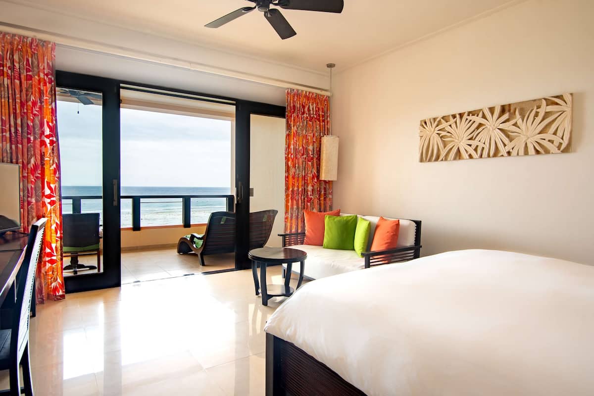 A hotel room with a bed, a desk, a chair, a small table, and a view of the ocean.