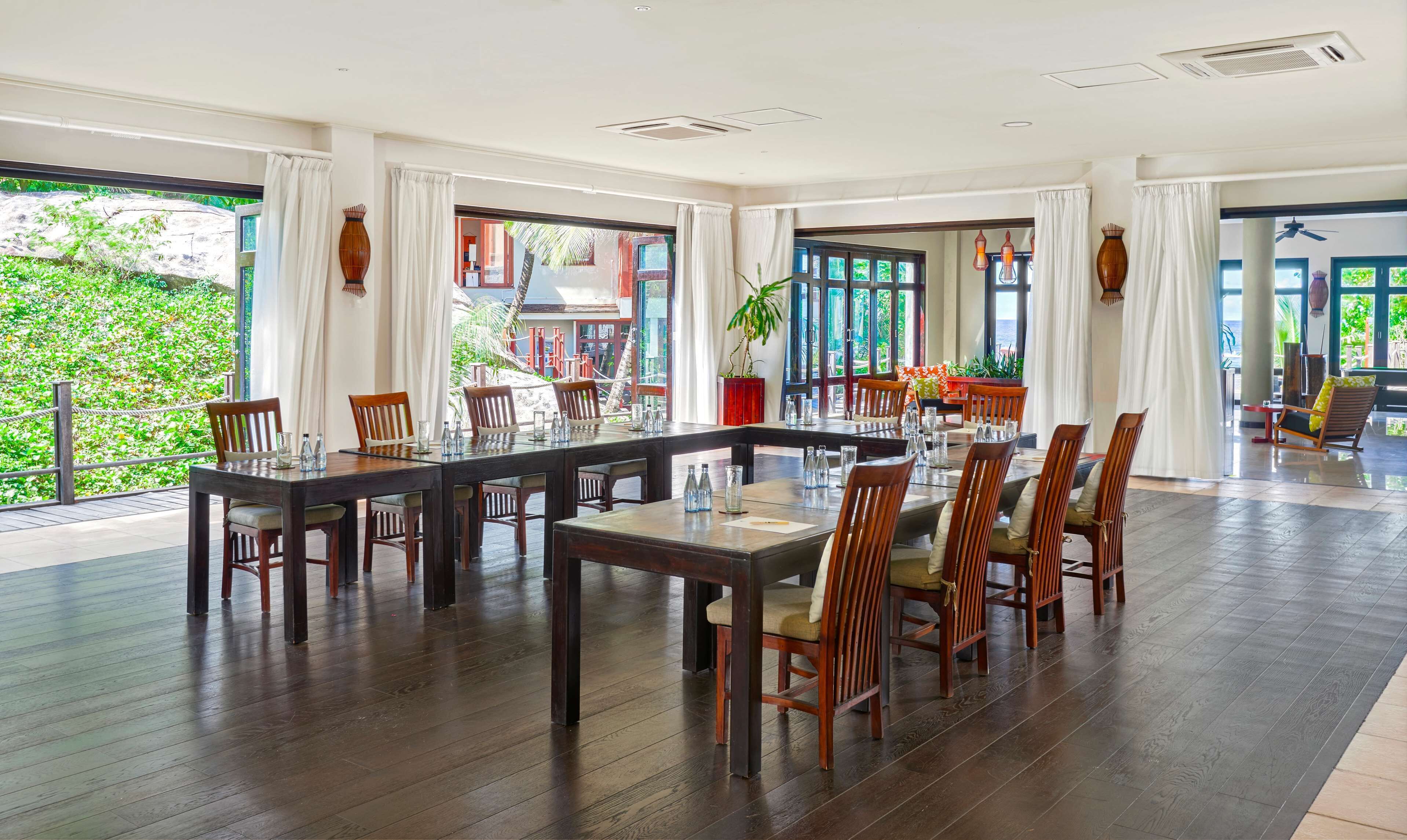 Une salle à manger spacieuse avec une grande table et des chaises, de grandes fenêtres avec des rideaux et une vue sur la verdure à l’extérieur.
