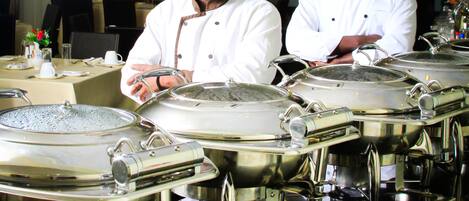 Dos chefs de pie detrás de una mesa de buffet con grandes recipientes de acero inoxidable.