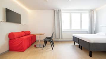 A modern hotel room with a bed, a red sofa, a small round table, and a TV mounted on the wall.