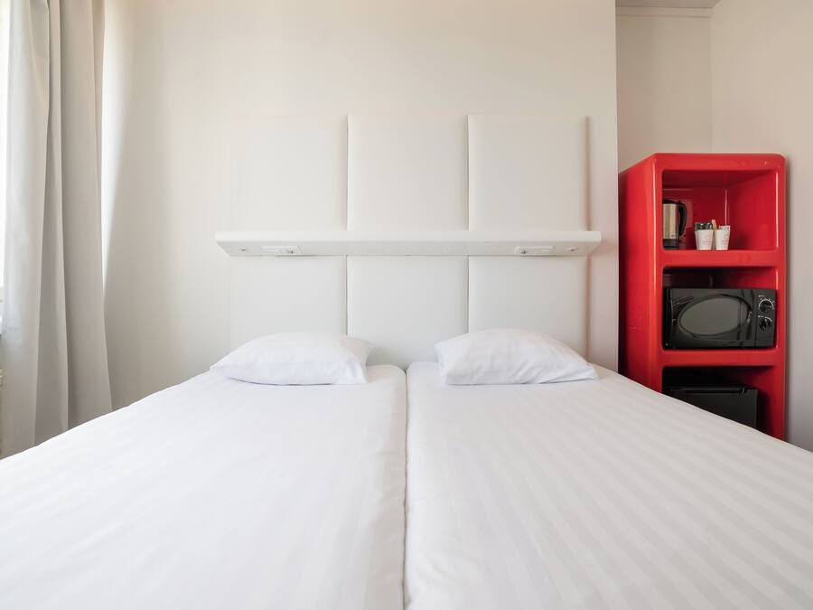 Una habitación de hotel con una cama, cortinas blancas, un estante rojo con un microondas y un cabecero blanco.