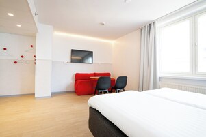 A modern hotel room with a large bed, a red sofa, two chairs, a TV mounted on the wall, and a window with curtains.