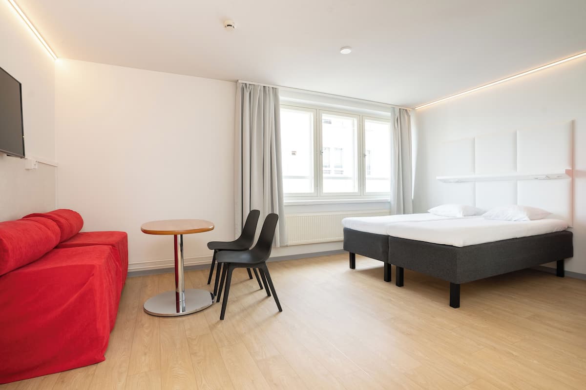 A modern hotel room with a bed, a red sofa, a small round table, and two black chairs.