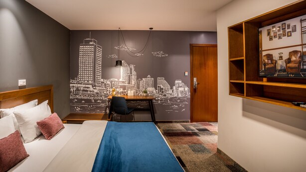 Une chambre d’hôtel comprenant un lit, un bureau, une chaise, une fresque murale représentant un paysage urbain et une bibliothèque.