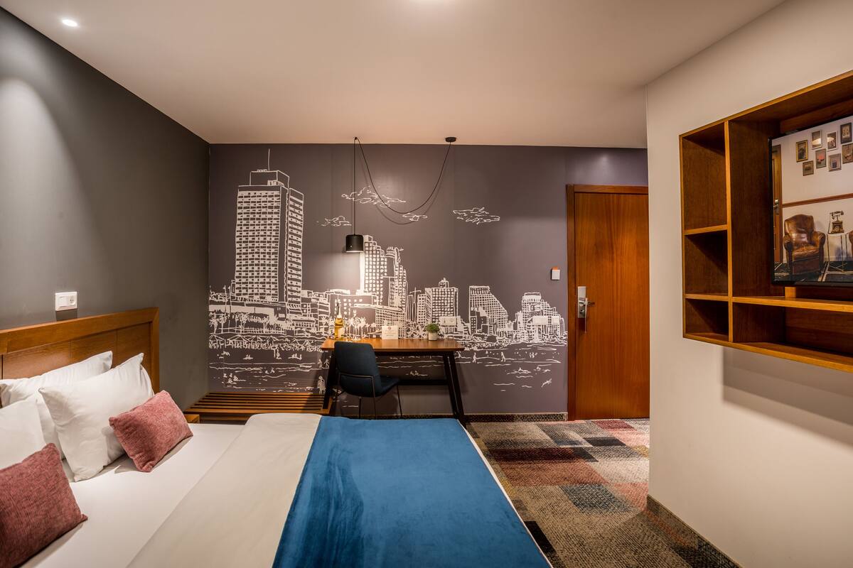 Habitación de hotel con un mural de paisaje urbano, una cama con almohadas blancas y rosas, un escritorio con una silla y un estante de madera con fotografías enmarcadas.
