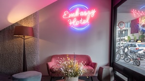 Un hall d'hôtel moderne avec une enseigne au néon indiquant « Embassy Hotel », un canapé rouge, une table ronde ornée d'un vase de fleurs et une vue sur les motos garées à l'extérieur.