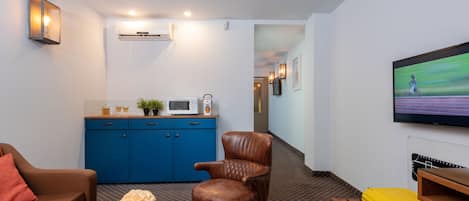 A compact living space with a brown leather chair, a blue cabinet with kitchen appliances, a small table with a vase of flowers, and a yellow suitcase on a bench.
