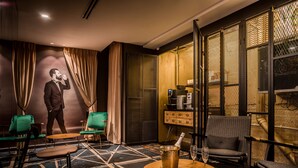 Un espace lounge moderne avec un sol à motifs géométriques, des fauteuils verts, une petite table ronde sur laquelle se trouvent une bouteille de champagne et un verre, et un grand écran mural qui affiche l’image d’un homme en costume.