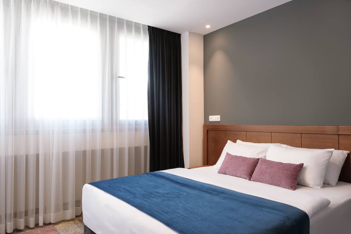 Habitación de hotel con una cama grande, cabecera de madera y cortinas translúcidas.