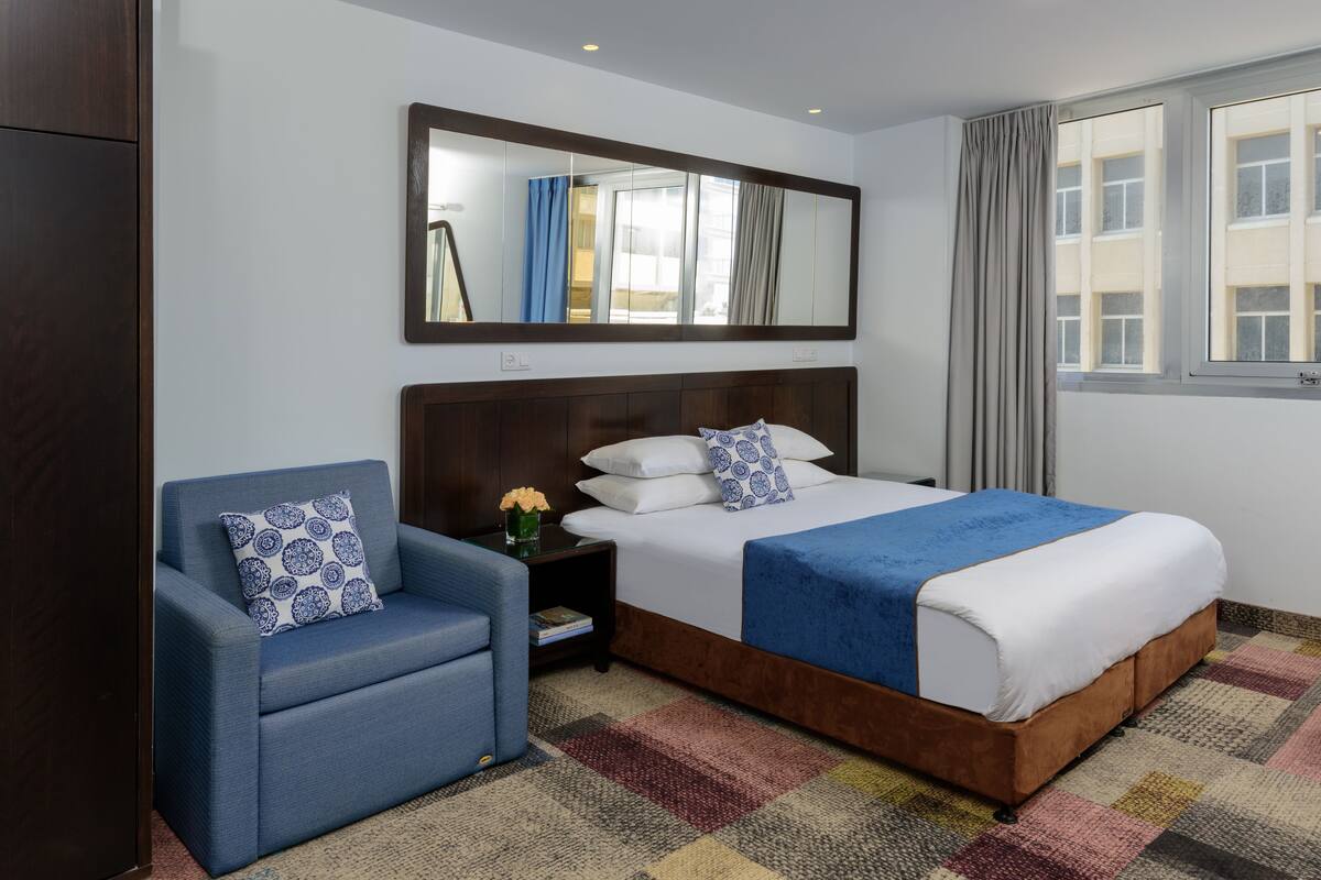 Una habitación de hotel con una cama grande, un sillón azul, una mesita de noche con un jarrón de flores, un espejo sobre la cama y una ventana con cortinas.
