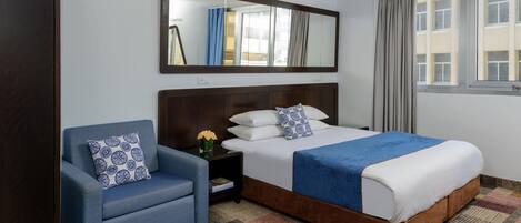 A hotel room with a large bed, a blue armchair, a nightstand with a vase of flowers, a mirror above the bed, and a window with curtains.