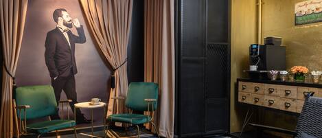 A hotel room with a large portrait, two green chairs, a small table, a coffee machine, and a cabinet with drawers.