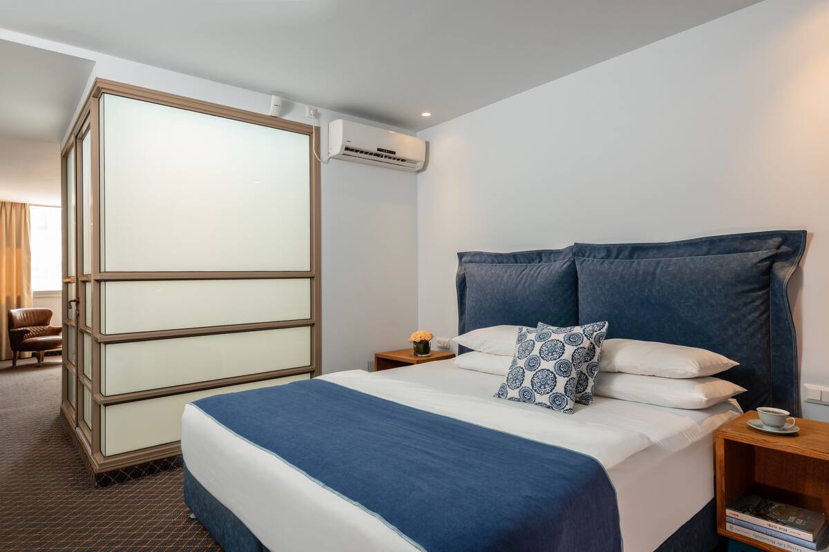Una habitación de hotel con una cama grande, una mesita de noche con lámpara, un aparador y un equipo de aire acondicionado.