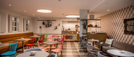 Una cafetería moderna con suelo de cuadros, varias zonas de estar que incluyen un sofá y mesas, y una diversidad de sillas.