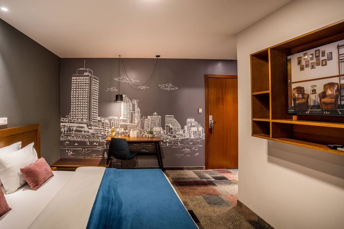 Una habitación de hotel con cama, escritorio, silla, un mural de paisaje urbano y una estantería.