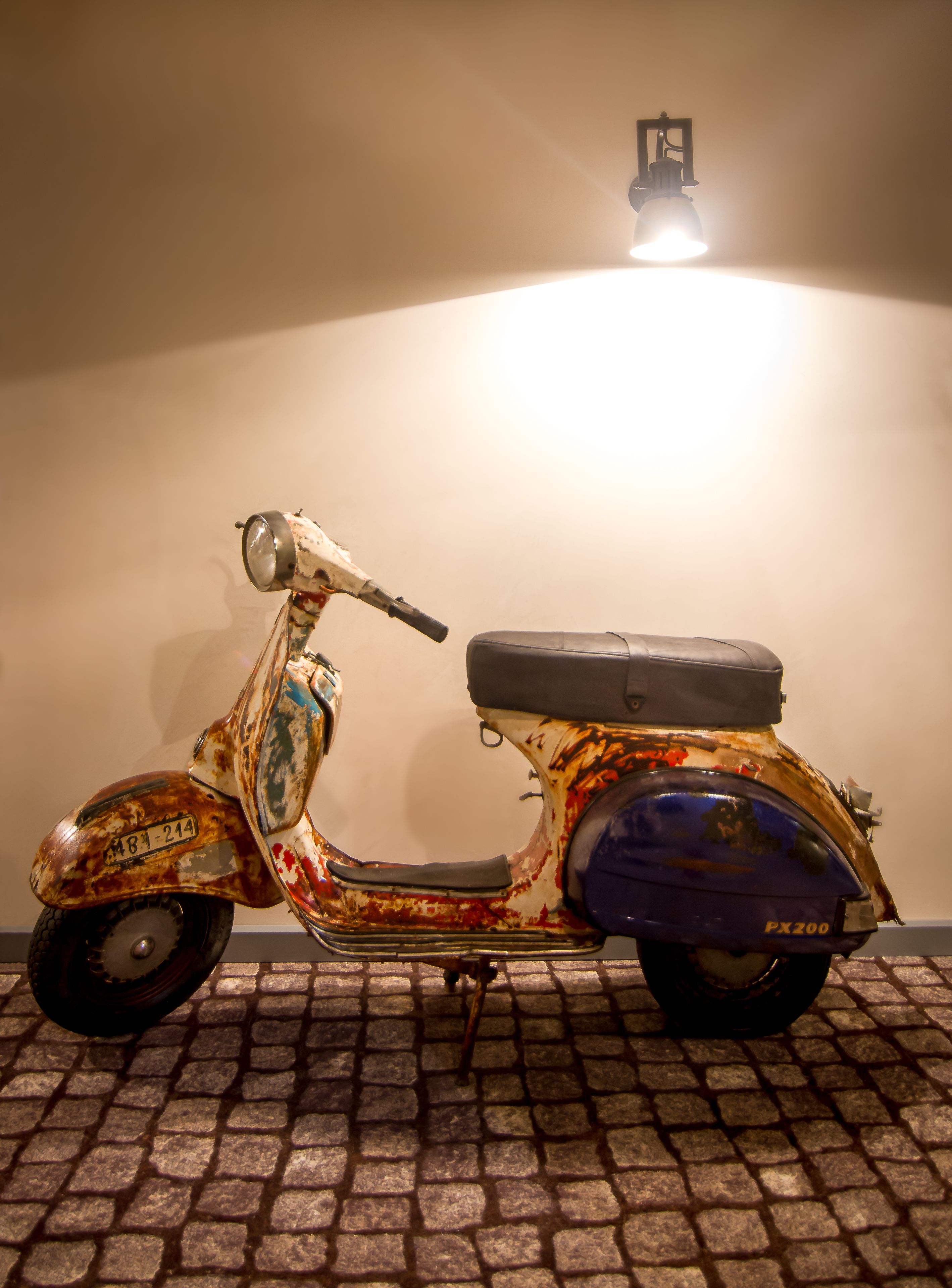 Un scooter vintage est garé sur une surface pavée, adossé à un mur sobre, avec un unique luminaire placé juste au-dessus.