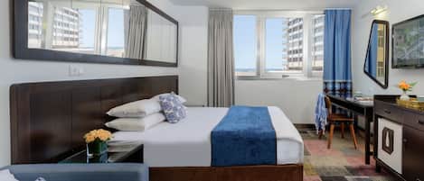 A hotel room with a large bed, a desk, a chair, a dresser, and a large window with curtains.