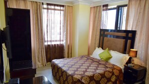 A hotel room with a bed, a television, a nightstand, and curtains. - New Avon Apartments (Dar es Salaam)