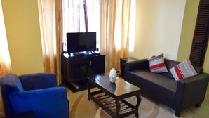 A living room with a blue armchair, a dark sofa with striped pillows, a coffee table with a vase, and a TV on a stand. - New Avon Apartments (Dar es Salaam)