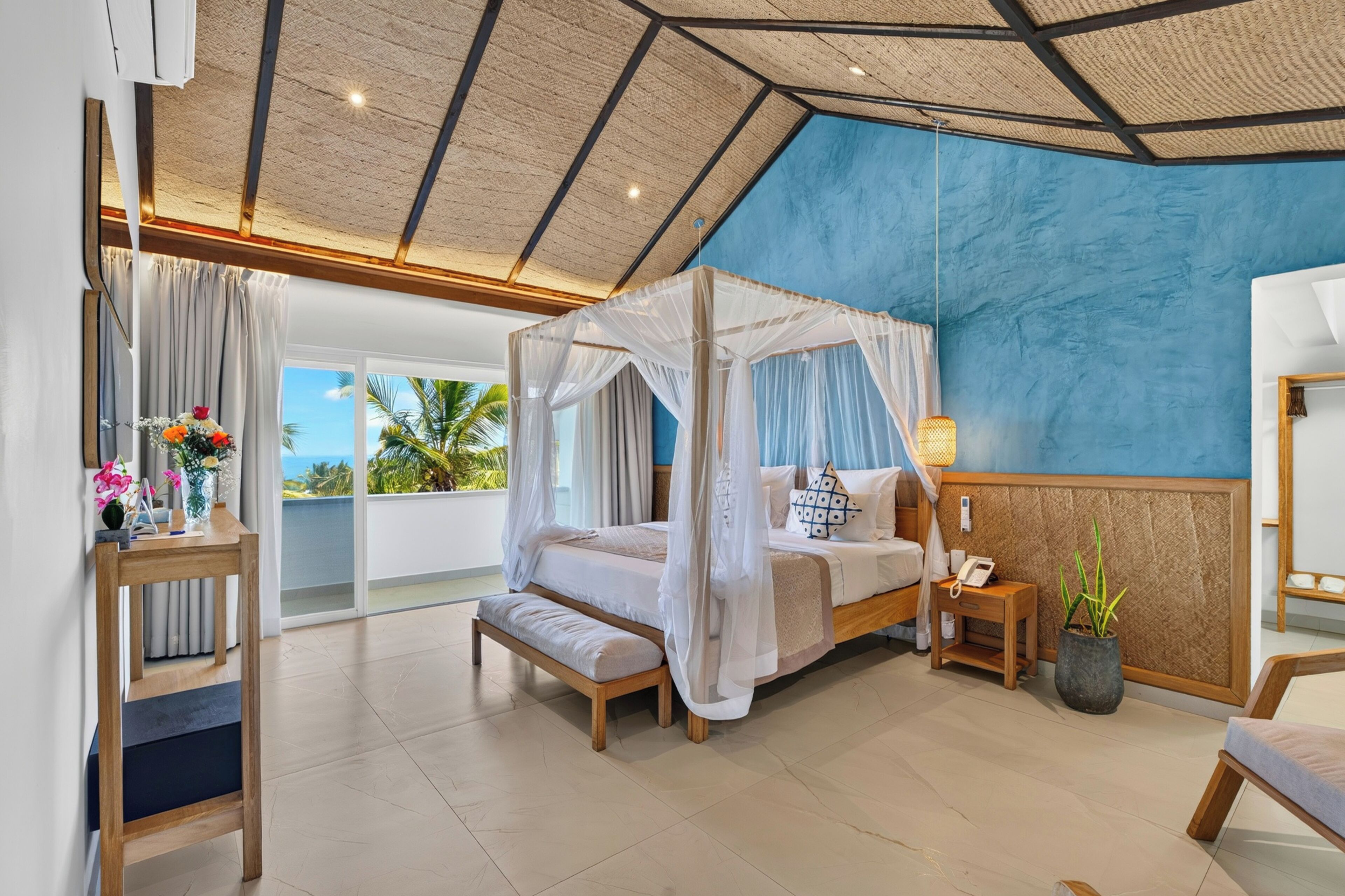 A bedroom with a four-poster bed, a bedside table with a lamp, a potted plant, and a view of the ocean through a large window.