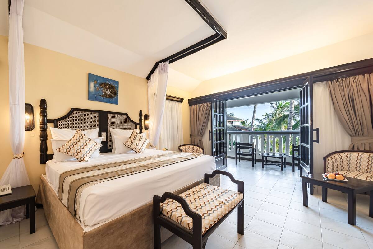 A hotel room with a bed, two chairs, a small table, and a balcony with a view of palm trees.