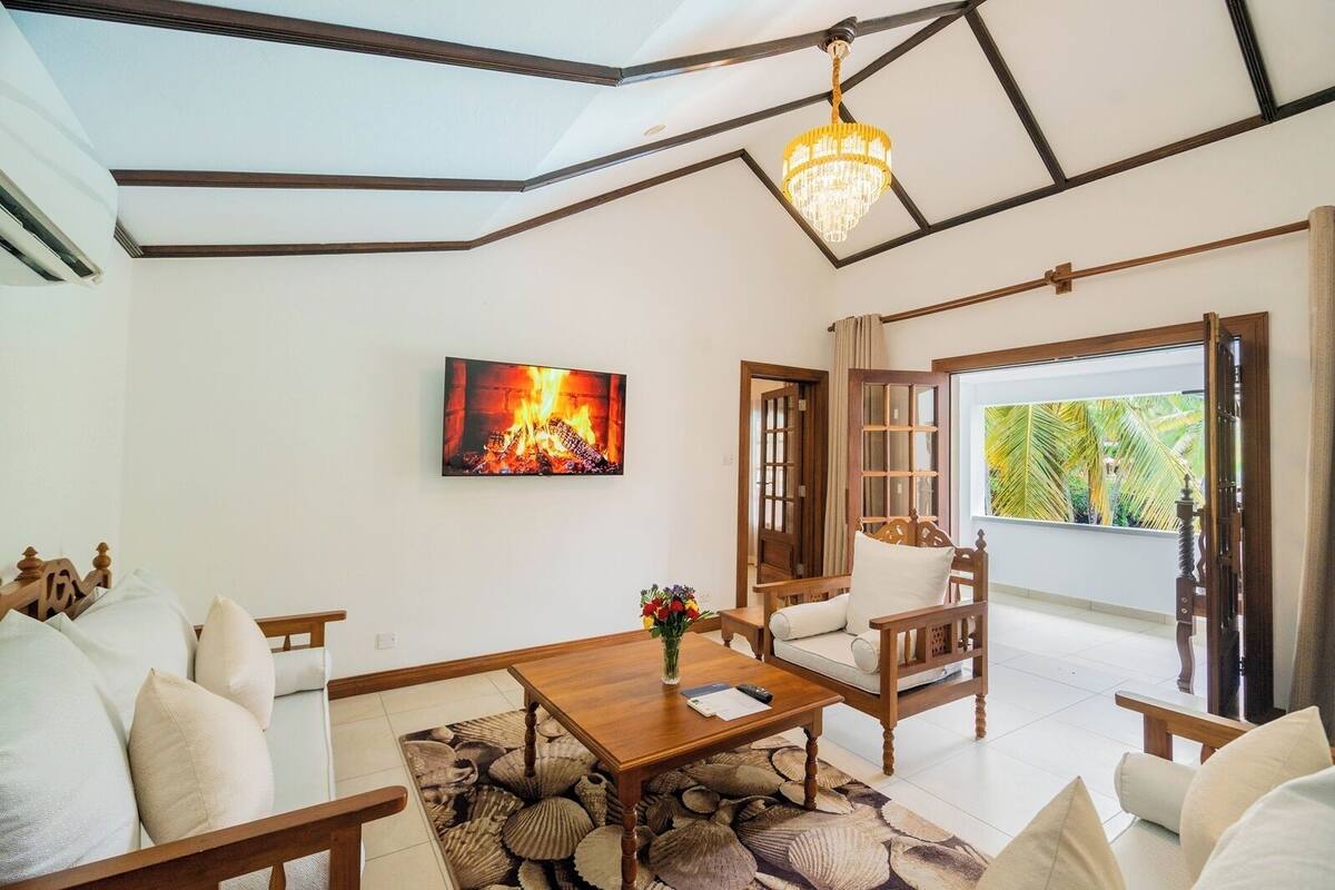 A spacious living room with a fireplace, wooden furniture, a chandelier, and a large window with a view of palm trees.