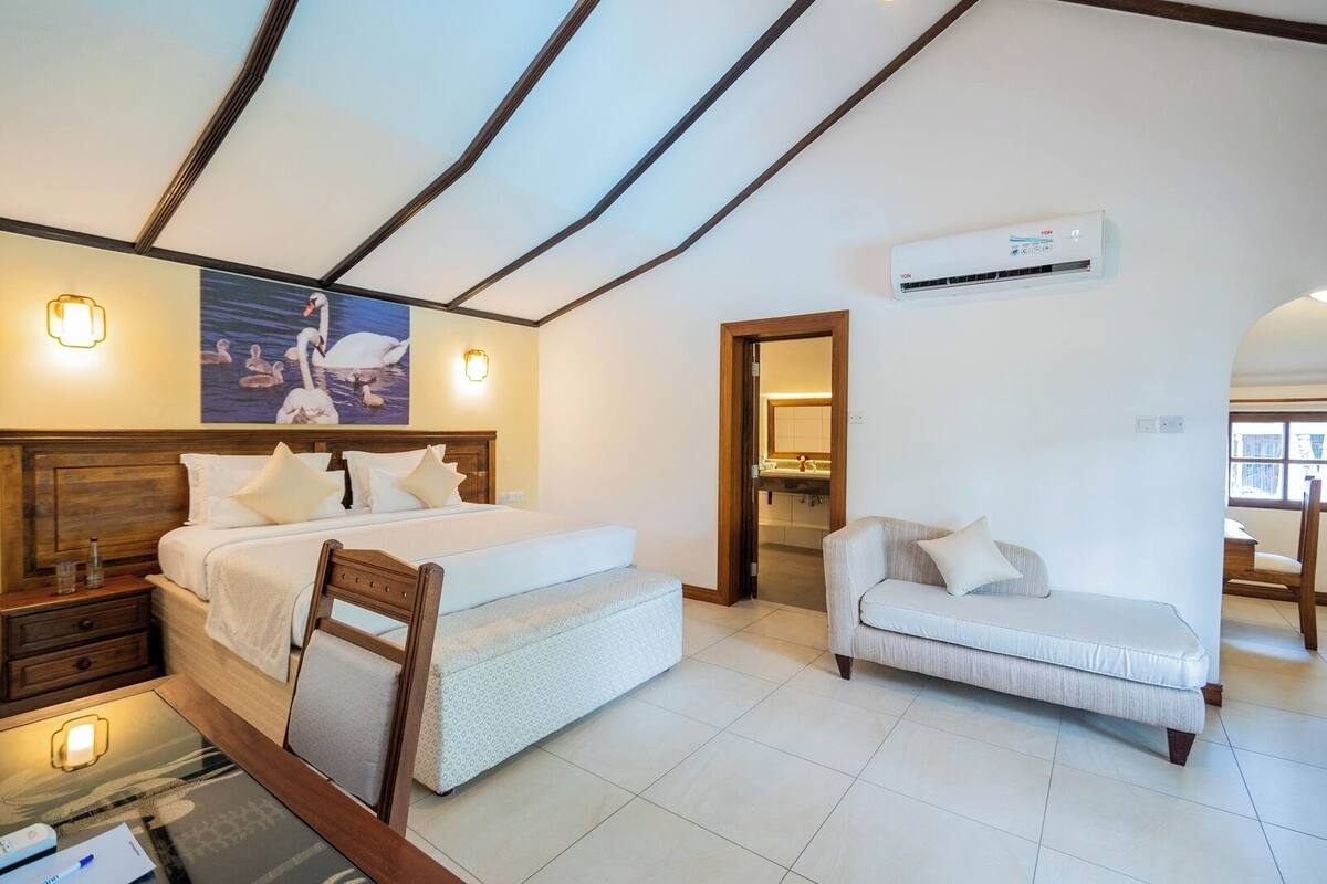 A bedroom with a bed, bedside tables, a chair, a sofa, a painting, and an air conditioning unit.