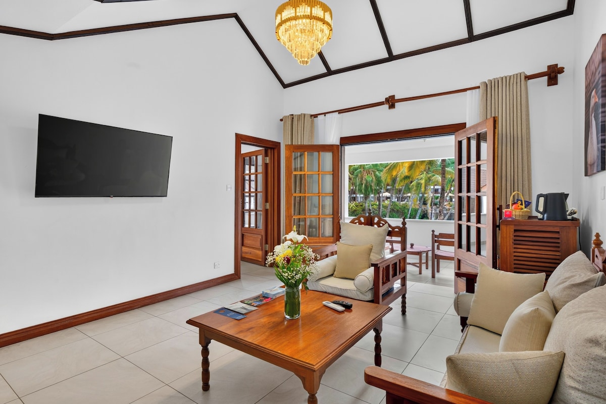 A modern living room with a flat-screen TV, a wooden coffee table, a sofa set, and a view of palm trees through an open door.