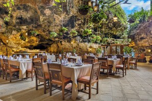 A restaurant with tables set for dining, a cave-like interior with hanging plants, and a rocky wall backdrop. - PrideInn Flamingo Beach Resort & Spa Mombasa (Mombasa)
