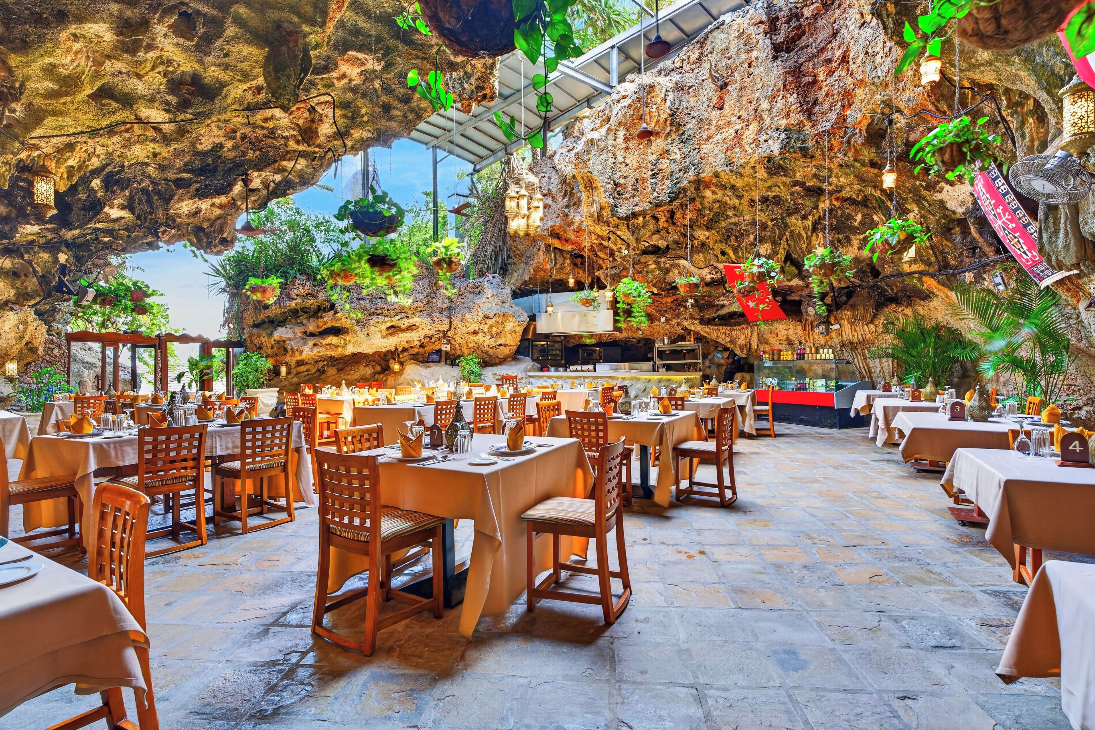 A restaurant with tables and chairs arranged in rows, a stone floor, and hanging plants.
