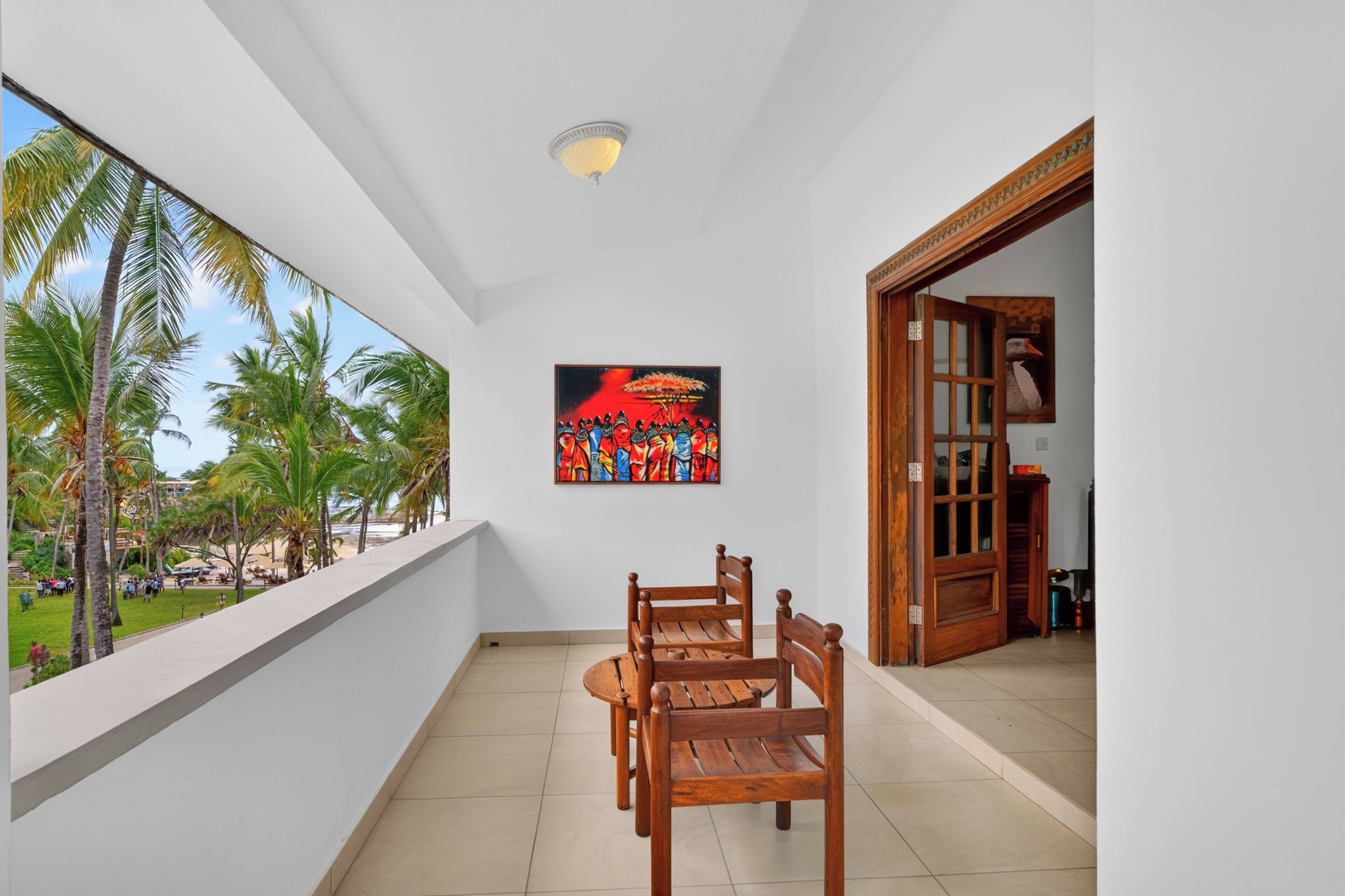 Uma varanda com mesa e cadeiras de madeira, um quadro na parede e vista para palmeiras e uma praia.