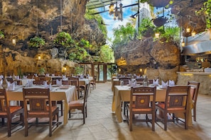 An indoor restaurant with tables set for dining, a kitchen area, and a view of the outdoors through a natural rock wall. - PrideInn Flamingo Beach Resort & Spa Mombasa (Mombasa)