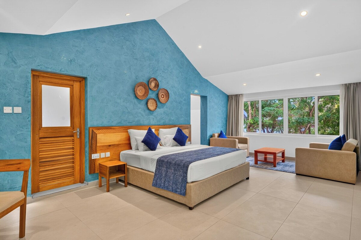 Une chambre moderne dotée d’un grand lit, d’une tête de lit en bois, d’un coin salon avec un canapé et des fauteuils, et offrant une vue sur la verdure environnante.