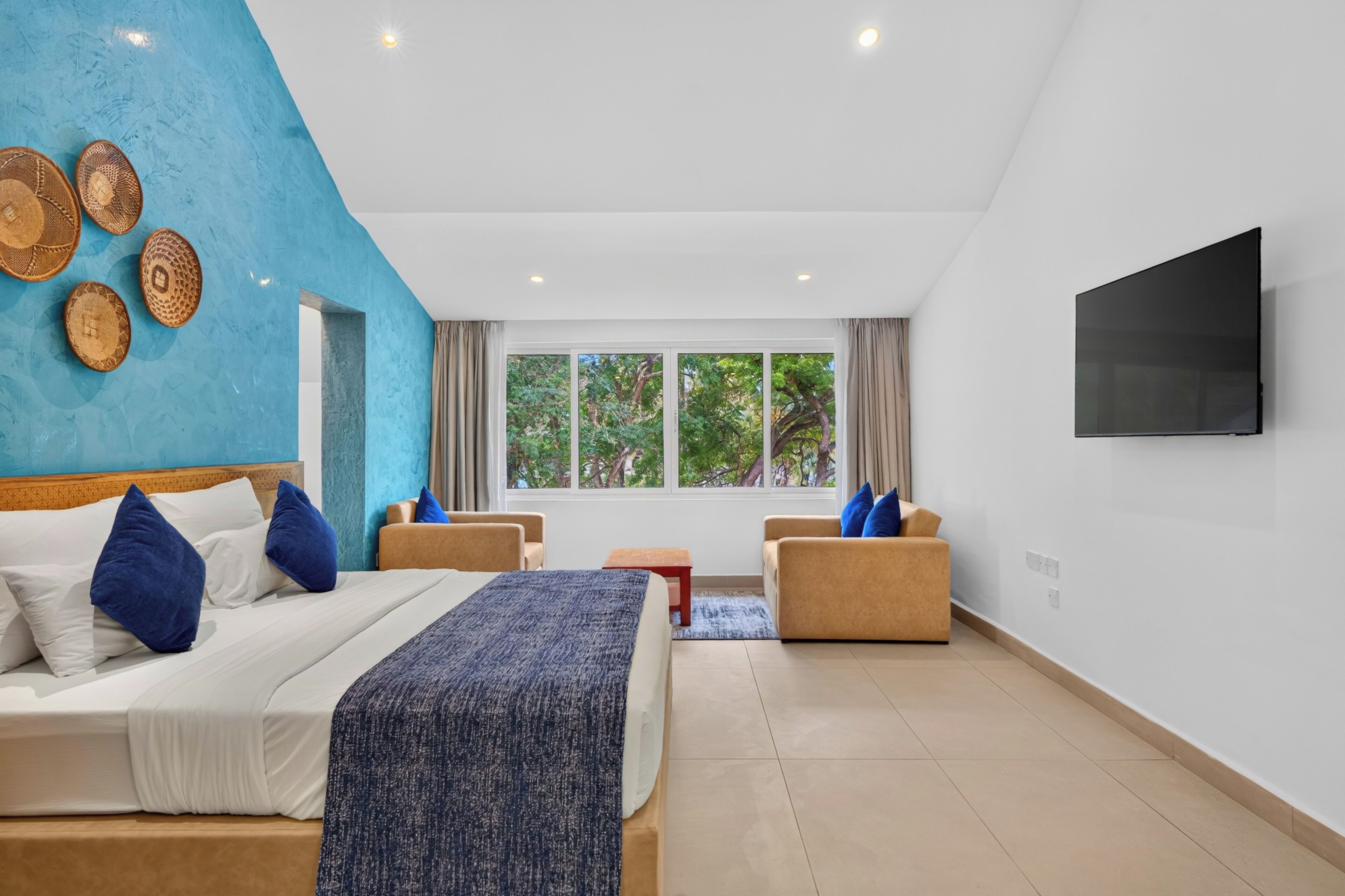 Habitación de hotel con cama, dos sillones, televisor y ventana grande con vistas a árboles.
