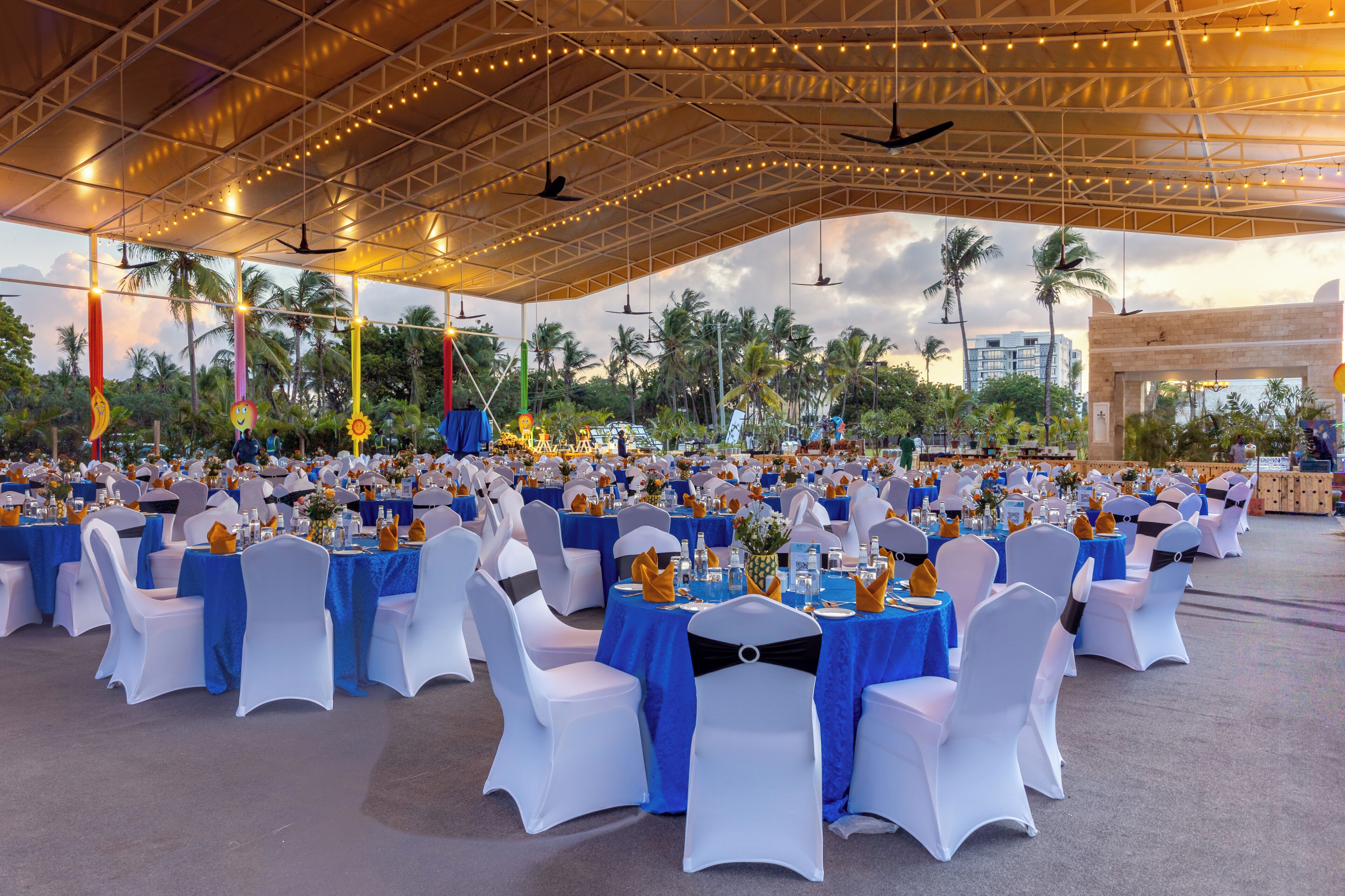 An outdoor event space with rows of tables and chairs, covered by a large canopy with string lights.