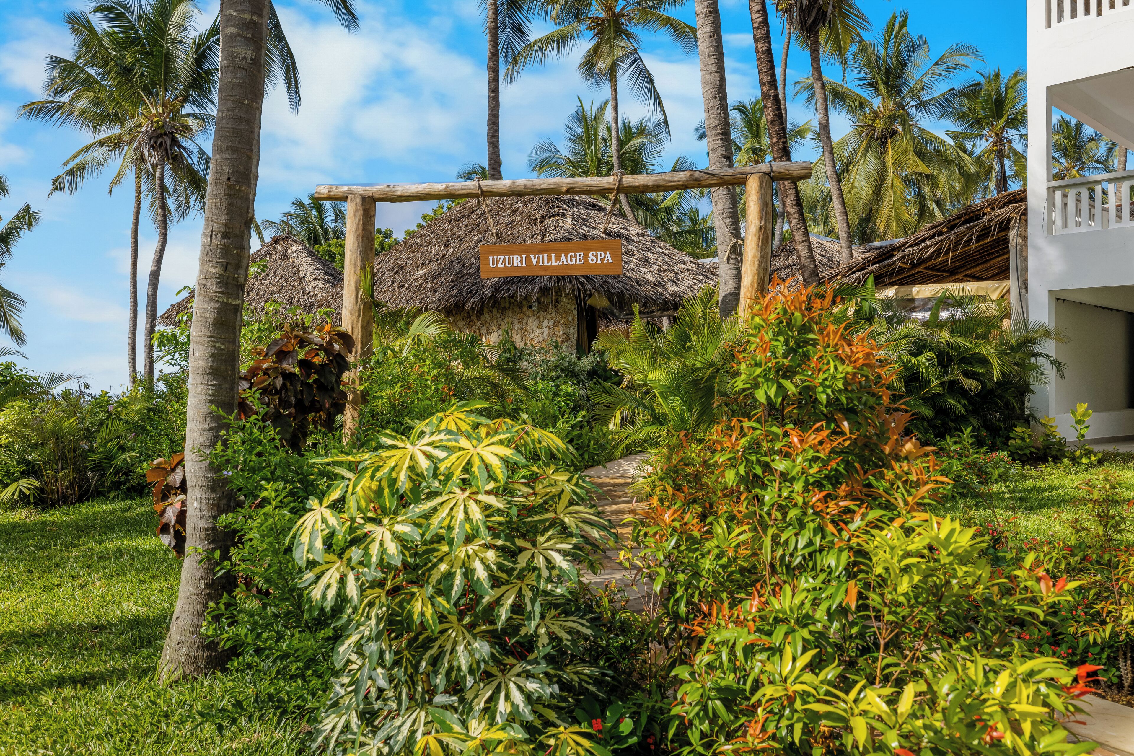 A thatched-roof entrance to Uzuri Village Spa surrounded by lush greenery and palm trees.
